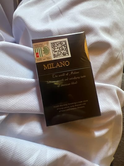 The image shows a pack of Milano cigarettes on a white textured fabric. The pack is predominantly black with gold lettering. On the top left of the pack, there is a design with green stripes, a red and white symbol, and a leaf. There is also a QR code on the top right. The text on the pack reads "MILANO" in large gold letters, followed by "The world of Milano" and "delivers smooth and satisfying taste of american blend." At the bottom of the pack, a health warning is printed, stating "Health Warning: Smoking is main cause of lung cancer, lung diseases and of heart and arteries diseases." The fabric appears to be soft and slightly wrinkled, creating shadows and highlights. There is no indication of the city of Machara, Abkhazia, or any people in the image.