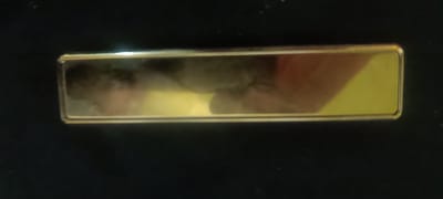 This is a close-up, top-down photograph of a rectangular, gold-colored object with a reflective surface. The object is positioned horizontally against a dark, matte background. The reflective surface shows a blurred, yellow-toned area on the right and a darker, cloudier area on the left, which appears to be a reflection of a person's hand and arm. There are no discernible landmarks or environmental cues to indicate a specific location in Semenivka, Ukraine. The lighting suggests an indoor environment with some ambient light sources causing glare on the reflective surface.