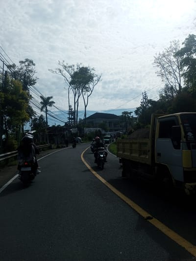 The image depicts a road in a rural or semi-rural area, possibly Khe Luang, Laos, based on the prompt, although it cannot be definitively confirmed. The road curves slightly and has a yellow line marking the center. Several motorcycles are visible on the road, both ahead and behind the viewer's perspective. A truck is parked on the right side of the road, partially obstructing the lane. The truck bed is filled with dirt or sand. Vegetation, including trees and shrubs, lines both sides of the road. Power lines are visible overhead, crisscrossing the sky. A building can be seen in the background, partially obscured by trees. The sky is overcast with scattered clouds.
