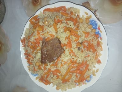 A close-up, overhead shot shows a plate of pilaf, a rice dish, centrally positioned on a white tablecloth with a floral pattern. The pilaf contains rice, thinly sliced carrots, and chickpeas, with a piece of cooked meat placed on top. The plate is white with a decorative blue floral rim. The background consists of the patterned tablecloth. There are no people visible in the image, and no specific location information, including the city of Tursunzoda, Tajikistan, can be determined from the visual content alone.
