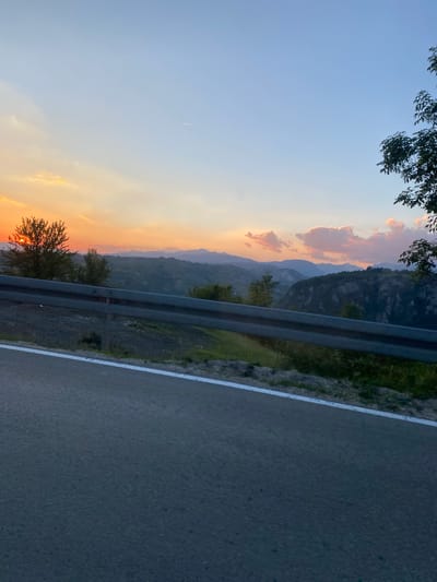 The image captures a scenic mountain landscape during sunset, viewed from a roadside. In the foreground, a paved road with a white line marking its edge stretches into the frame. A metal guardrail runs along the side of the road, separating it from a sloped embankment covered in grass and some scattered rocks. Beyond the guardrail, the terrain descends into a valley characterized by rolling hills and dense vegetation. Further in the distance, layered mountain ranges fade into the horizon, under a sky painted with warm hues of orange, yellow, and pink, indicative of the setting sun. A few wispy clouds are present, and the sun itself is partially visible, peeking through a tree on the left side of the image. The overall scene suggests a natural, undeveloped environment, likely in a rural mountainous region like Šavnik, Montenegro. There are no people or visible signs of urban development. The time of day is clearly sunset.