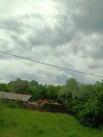 The image depicts an outdoor scene with a cloudy sky dominating the upper portion. Below the sky, a dark, thin wire stretches horizontally across the frame. In the middle ground, a dense line of green trees forms a lush backdrop. A low-lying structure with a light-colored roof and exposed wooden beams is partially visible on the left, situated behind a dark fence. To the right of this structure, there appears to be a red object, possibly part of a vehicle or equipment, and a small gazebo. The foreground consists of a bright green, grassy slope that appears to be moving or blurred, suggesting the photo was taken from a moving vehicle. The overall impression is a rural or semi-rural setting, possibly on the outskirts of Saratovskaya, Russia, under overcast conditions.