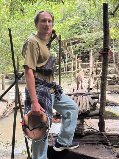 A man stands in front of a natural scene in Ban Huai Takhrai, Thailand. He is wearing a tan t-shirt, a blue and black plaid shirt tied around his waist, light blue jeans, and dark sneakers. He holds a brown and black motorcycle helmet in his right hand. A small brown dog stands on a wooden bridge in the background, facing away from the camera. The environment consists of lush green trees and foliage, with a body of water flowing through rocks. A rustic wooden structure, possibly a bridge or walkway, is visible in the mid-ground. The lighting suggests it is daytime, with diffused natural light.