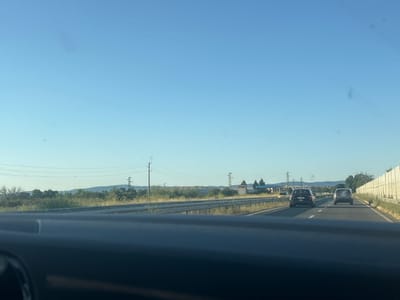 The image captures a view from inside a vehicle traveling on a highway. The sky is a clear, light blue, suggesting a sunny day. The highway stretches ahead, with two lanes of traffic visible. Several cars are on the road, including two dark-colored SUVs in the right lane and a yellow car further ahead. The median barrier separates the lanes, and on the right side of the highway, a light-colored concrete sound barrier is present.

The surrounding landscape is a mix of dry, grassy vegetation and scattered trees, with rolling hills in the distance. Power lines and utility poles are visible along the side of the road, indicating infrastructure in the area. In the background, there are some buildings and structures, possibly indicating a small town or industrial area. One building appears to be a small white structure with a tower.

The overall scene conveys a sense of travel and movement on a highway. The time of day appears to be late morning or early afternoon, given the brightness of the sky and the quality of the light. There are no people directly visible in the image, but the presence of cars implies human activity. The location context provided is Aheloy, Bulgaria. There is no discernible text in the image.