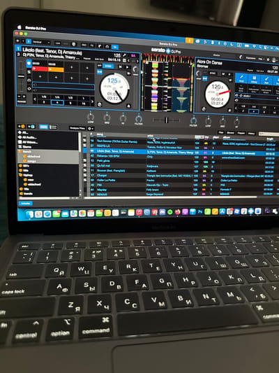 The image shows a gray MacBook Air laptop open, displaying the Serato DJ Pro software. The screen is angled slightly to the left, and the keyboard, which features Cyrillic characters alongside Latin letters, is partially visible. The Serato software displays various waveforms, track information, and DJ controls, including tempo, track names, and EQ settings. Icons for other applications, such as Safari, Mail, Messages, and Spotify, are visible in the macOS dock. There are no people visible. The location is not identifiable from the image.