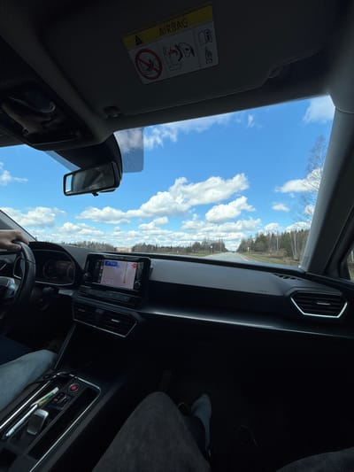 The image, taken from the interior of a car, captures a bright daytime scene looking out through the windshield. The perspective is from the driver's side, with a hand visible on the steering wheel.
Outside the car, a wide expanse of clear blue sky with scattered fluffy white cumulus clouds fills the upper and central parts of the frame. A road, likely paved, stretches into the distance, flanked by dense forest composed mainly of bare deciduous trees, suggesting early spring or late autumn.
Inside the car, various elements of a modern vehicle interior are visible. The dashboard includes a central infotainment screen displaying a navigation map, with some numbers like "118" and "173" faintly visible. To the right of the screen, an air vent with a glowing outline adds a modern touch. Further right, a foot wearing a white sock is visible in the passenger footwell. The rearview mirror is present, reflecting a partial view of the interior.
Above, the sun visor is lowered, featuring a yellow warning label with the prominent text "AIRBAG" and universal icons depicting child seat and airbag safety information. A smaller "AIRBAG" text is also discernible near the passenger-side air vent. Below the infotainment system, controls for the climate and other car functions are partially visible, including a red button. The scene depicts a journey in progress along a road in what is identified as Печмень, Russia, under clear and sunny weather conditions.