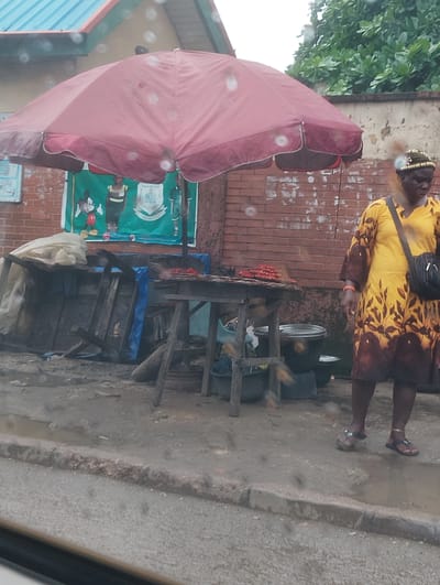 The image depicts an outdoor scene in Alagbede, Nigeria, with a woman standing to the right of a food stall. The woman is wearing a yellow and brown patterned dress and a dark shoulder bag. She has on sandals and jewelry on her wrist and ankle. The food stall features a red umbrella, a wooden table with food items on it, and various containers. Behind the stall, there is a brick wall and a banner with cartoon characters and text. The ground is wet and appears to be a street or pathway. Trees are visible in the background. The overall impression is of a casual street scene, possibly in a market or roadside area.