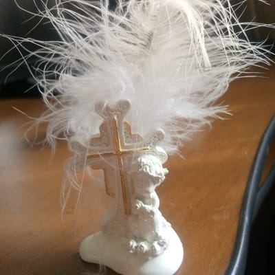 A close-up, slightly angled shot captures a small white ceramic angel figurine with gold accents, standing on a light brown wooden surface. The figurine is adorned with a spray of white feathers, creating a halo-like effect. The angel appears to be holding something in its arms. The background is dark and out of focus, suggesting an indoor setting. The overall impression is one of delicate ornamentation. This scene is not located in Ashtarak, Armenia.