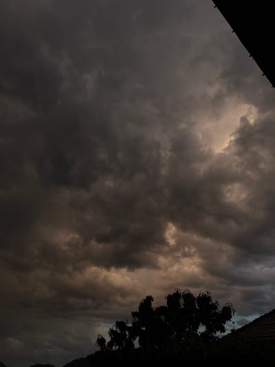 The image shows a sky filled with dark, ominous clouds, suggesting an impending storm. A silhouette of a tree and building are located on the right. A corner of a dark roof is visible in the top left corner. The location is possibly Zhaltusha, Bulgaria.