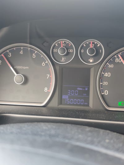 The image shows a close-up of a car's dashboard. The tachometer reads around 2200 rpm. The engine temperature gauge shows a reading around 50 degrees Celcius. The fuel gauge reads "full". The digital odometer displays 150000 km and the trip meter displays 300 km. The speedometer indicates that the car is traveling at approximately 80 km/h. There is a green light illuminated in the lower right of the speedometer. The steering wheel is visible in the lower portion of the frame. There is no information available to connect this image to Jeleznitsa, Bulgaria.