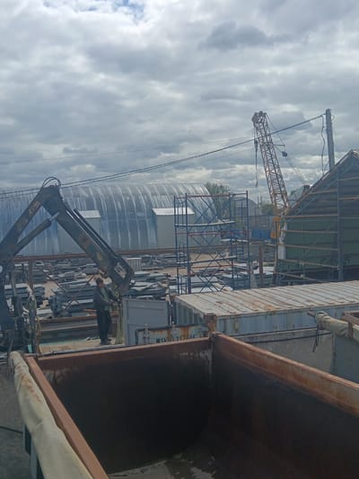 The image, taken in Яльгелево, Russia, shows a construction or industrial yard on an overcast day. A person wearing dark clothing stands on a flatbed trailer, appearing to operate a crane that is lifting a metal panel or plate. The crane is positioned next to a dumpster or large container. In the background, there are stacks of metal plates or tiles, scaffolding, shipping containers, and a large, corrugated metal structure that resembles a hangar. A yellow construction crane is visible further in the background. Power lines are strung across the sky.