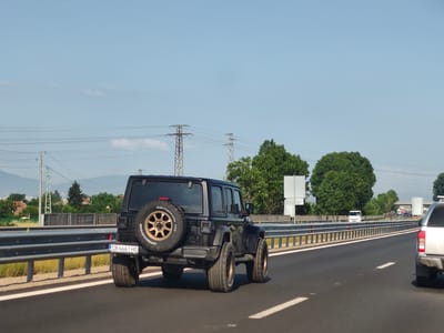 In Gorni Bogrov, Bulgaria, on a clear day with a light blue sky, a black Jeep Wrangler with a spare tire mounted on the back drives on a highway. The Jeep's license plate reads "CB 6661 HO". The highway has a metal guardrail separating the lanes. In the background, there are green trees, houses, power lines, and distant mountains. Another vehicle, a white pickup truck, is visible in the right lane ahead of the Jeep.