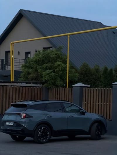 A grey Kia Sportage is parked on a street in front of a modern house with a dark grey metal roof and light-colored brick walls. A yellow pipe runs horizontally across the scene in front of the house. A wooden fence is in the foreground, partially obscuring the car. The sky is overcast and a pale blue. The license plate of the car is visible and reads "560 INA". The image appears to be taken in the city of Orto-Sai, Kyrgyzstan.