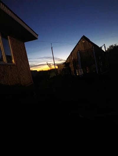 The image shows an outdoor scene at dusk, with a deep blue sky transitioning to lighter hues near the horizon. In the foreground on the left, a part of a building with a brown, textured facade and a sloped roof is visible. A window with yellow trim reflects the sky. To the right, a wooden structure, possibly a shed or a small house, stands with a gabled roof. Its dark silhouette is contrasted against the fading light. Power lines and a utility pole are also present in the background. The ground is dark and appears to be covered in vegetation. The overall setting suggests a rural or semi-rural environment. The location is identified as Buzov'yazy, Russia.