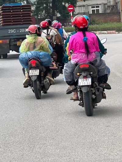 Two individuals are riding on the back of a motorcycle, traveling on a paved road in Phúc Yên, Vietnam. The motorcycle is dark-colored with a visible license plate reading "18-H1 710.92". The passenger in the foreground is wearing a bright pink long-sleeved shirt and dark pants with a white polka dot pattern. They have dark hair braided and tied with a blue scrunchie. They are also wearing a clear plastic poncho over their clothing. The driver of the motorcycle, partially obscured, is wearing a dark green jacket. In the background, another motorcycle is visible with two occupants. The person in the front wears a bright yellow translucent poncho over a pink garment, and their license plate reads "23-P1 103.41". They have dark hair and are wearing blue pants. The environment appears to be a street in an urban or semi-urban area, with buildings and trees visible in the distance. A large truck with wooden planks stacked on its bed is also present in the background, with a license plate starting with "29C". The road surface is asphalt.