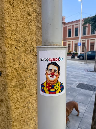 The image shows a sticker on a lamppost in La Línea De La Concepción, Spain. The sticker features a cartoon drawing of a man with clown makeup, including a red nose and a colorful outfit with a large bowtie. The text "EuropayasosSur" is printed above the man's head. The background of the image shows a textured yellow wall, a sidewalk, a building with red and white stripes, a parking sign, a scooter, a car, and a dog walking on the sidewalk. The sky is a light blue, suggesting it is either early morning or late afternoon.