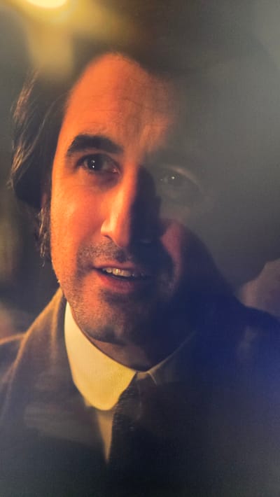 A close-up, slightly out-of-focus shot shows the face of a man wearing a suit and a light-colored shirt with a high collar. The man has dark hair that is parted and falls to the sides of his face. He has dark eyebrows, brown eyes, and a slight smile. The lighting casts a warm glow on his face, with shadows obscuring parts of it. The background is dark and indistinct. There is no discernible information about the location in Vlaardingen, Netherlands.