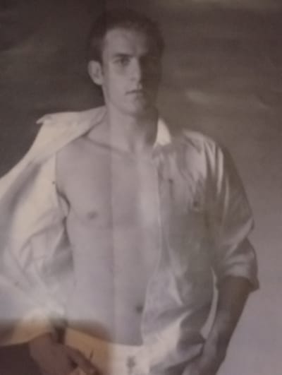 The black and white, slightly blurred image shows a young man in an open white shirt, revealing his bare chest. He is looking towards the camera, with his right arm bent and his hand resting on what appears to be his hip or thigh area, partially obscured by the shirt. His left arm is also bent, with his hand possibly reaching towards the shirt buttons. The background is a muted, dark tone, suggesting an indoor setting with soft lighting. The man's hair is dark and neatly styled. The image evokes a sense of casualness or vulnerability. There are no discernible elements that would specifically indicate the location within Berezivka, Ukraine.
