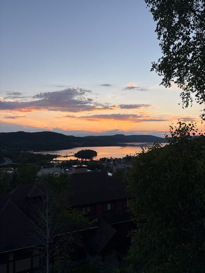 This is an aerial, eye-level, medium shot of a sunset over the city of Mont-Tremblant, Canada, viewed from a vantage point with foreground trees and rooftops. The sky is a gradient of soft blues and oranges, with wispy clouds catching the last rays of the sun. In the mid-ground, a large body of water, likely Lac Tremblant, reflects the colorful sky, with a small island dotted with buildings in the center. The distant shore is lined with rolling, tree-covered hills. A town with a cluster of buildings, possibly the ski village, is visible nestled near the water's edge. In the foreground, the dark, angled roofs of several buildings are prominent, along with the silhouetted branches of trees. A parking lot with several cars is visible in the middle ground to the left of the town. There are no people visible in the image.