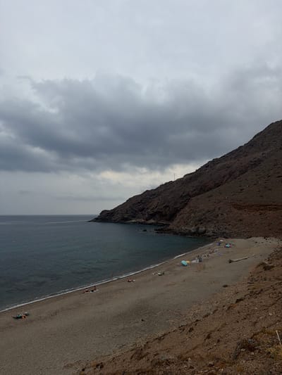 This is a tilted, high-angle shot of a pebble beach next to a rocky cliff face under an overcast sky. Several people are scattered along the beach, some lying on towels or blankets, others sitting or walking. Beach umbrellas, one yellow and one blue, are visible. A piece of driftwood lies on the sand. The sea is a dark, choppy grey. The clouds are heavy and dark, suggesting a moody, perhaps approaching or recent, rain. The lighting is dim, indicating it's likely late afternoon or a heavily overcast day. The rocky cliff is rough and uneven, with sparse vegetation. The sand is a mixture of small pebbles and darker grains.