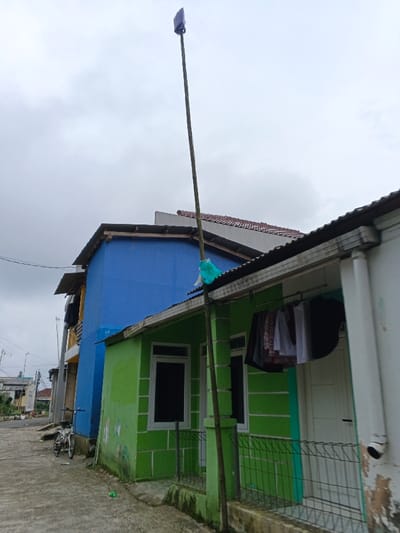 The image portrays an outdoor residential area in Kabiufa, Papua New Guinea, under an overcast, light-grey sky. A paved path occupies the lower left quadrant, where a white bicycle with dark tires is parked, leaning against a building. In the midground, two distinct houses are visible. The structure on the left is a two-story building painted bright blue, with exposed wooden rafters supporting its flat roof. Immediately to its right is a single-story house painted lime green, featuring a corrugated metal roof and a small metal fence across its front porch. This green house has two windows with dark panes and white trim, and a white front door. Green algae or mildew stains are present on the lower portion of its exterior walls. A collection of assorted clothes hangs on a rod beneath the green house's roof overhang. A tall, slender pole, appearing to be wood or bamboo, rises vertically from the ground in front of the green house, secured to its roof overhang with a light-green material, possibly plastic or fabric. A dark, rectangular antenna is affixed to the very top of this pole. To the far right, a partial view of an adjacent, lighter-colored building with a vertical downspout shows areas of peeling paint. Overhead utility wires are visible in the distant left background. No individuals are present in the scene.