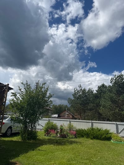 The image depicts a suburban outdoor scene under a dramatic sky with both dark storm clouds and bright blue sky visible. In the foreground, a lush green lawn is partially shaded. To the left, a white car is parked near a wooden structure with a thatched roof. In the midground, a small tree with green foliage stands to the left of a silver corrugated metal fence. Flowerbeds with pink and red flowers are situated in front of the fence. Behind the fence, there is a residential building with a brown roof and a treeline of evergreen trees. Power lines are visible across the upper portion of the scene. The location is identified as Bolshoe Kozino, Russia.