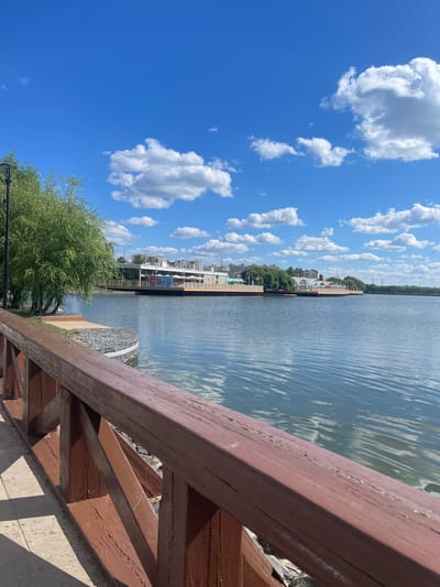 The image depicts a sunny day with a blue sky filled with white clouds. In the foreground, a wooden railing, painted brown, runs diagonally across the frame. Beyond the railing is a paved walkway and a body of water, likely a lake or river, with ripples reflecting the sky. Across the water, a long wooden pier or boardwalk extends, lined with buildings that appear to be restaurants or cafes with white awnings. Trees are visible behind the buildings, and in the distance, several apartment buildings suggest an urban setting. The scene is tranquil and appears to be a public park or recreational area. This location is likely in Moscow, Russia, given the architectural style of the buildings and the presence of such waterfront recreational spaces. There are no people visible in the image.