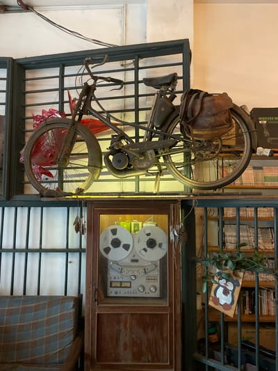 The image shows a vintage moped suspended on a dark metal grate in what appears to be a cafe or shop in Lien Nghia, Vietnam. The moped is dark in color with a worn leather seat and a leather saddlebag on the rear. A red object, possibly a paper lantern or decoration, is visible attached to the front wheel. Below the moped, a wooden cabinet with glass doors displays a vintage reel-to-reel tape recorder. The background reveals shelves filled with books and other miscellaneous items. A plaid-patterned sofa is visible in the lower left corner. The overall ambiance suggests a retro or vintage-themed establishment.