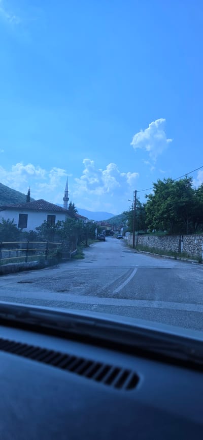 The image depicts a rural street scene in Melivoia, Greece, captured from the interior of a vehicle, evidenced by a dark dashboard with vents at the bottom of the frame. A paved road, appearing worn with some visible patches, stretches into the distance, with a single white road marking partially visible in the center.
On the left side of the road, a traditional white building with a terracotta tiled roof and dark-framed windows is prominent. Behind this building, a slender, light-colored minaret with a pointed spire rises into the sky, indicating the presence of a mosque. Green trees and shrubbery are visible around the building and along a wooden fence lining the road. Steep, tree-covered mountains form the immediate backdrop on the left.
On the right side of the road, a dry-stone wall extends, topped with additional fencing, with lush green trees behind it. A utility pole with electrical wires stands near the wall. In the distance, other buildings and further mountains are discernible. The sky is bright blue with scattered white, cumulus clouds. No individuals are visible in the scene.