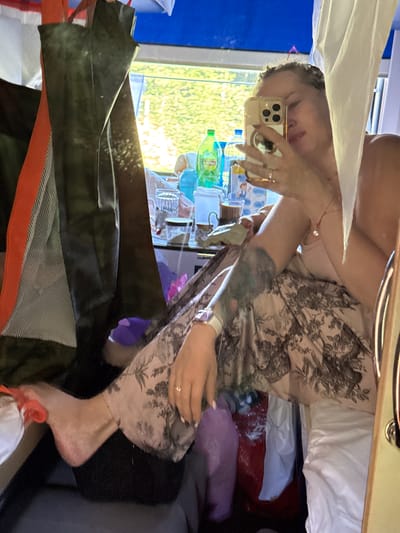 The image shows a woman taking a selfie in a narrow interior space, possibly a train compartment or a small cabin. She is sitting with her legs crossed, wearing patterned trousers and a light-colored top. Her left arm is extended, holding a smartphone. A large tattoo is visible on her left forearm. The background is cluttered with various items, including bags, bottles, and containers. Sunlight streams in from a window behind her, illuminating some of the objects and creating a somewhat dim environment. The scene appears to be inside a moving vehicle, given the reflection of greenery outside the window. The image does not provide any specific visual cues that would allow for its localization to Stupino, Russia, or any other specific city.