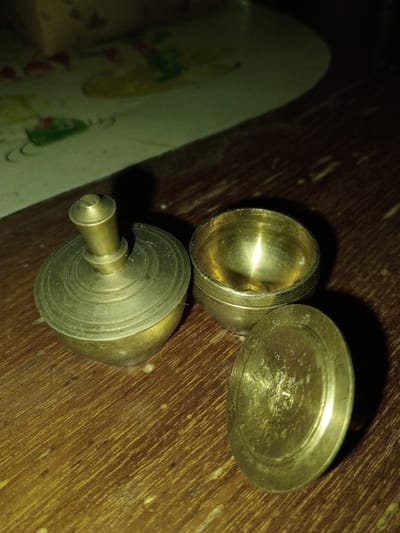 The scene depicts three individual brass items arranged on a dark, wooden surface in Longmiang, Laos. To the left, a lidded brass vessel with a central finial sits upright, its upper surface adorned with concentric rings. Adjacent to it, a small, open brass container with a rounded bottom displays an empty, tarnished interior. To the right, a circular brass lid, likely corresponding to the open container, is positioned vertically on its edge, also exhibiting concentric rings on its surface. The wooden surface displays a visible grain pattern. The background is dark and out of focus, with a light-colored, patterned mat partially visible in the upper left. A direct light source illuminates the brass objects from the upper left, creating distinct reflections and shadows. No individuals are present.