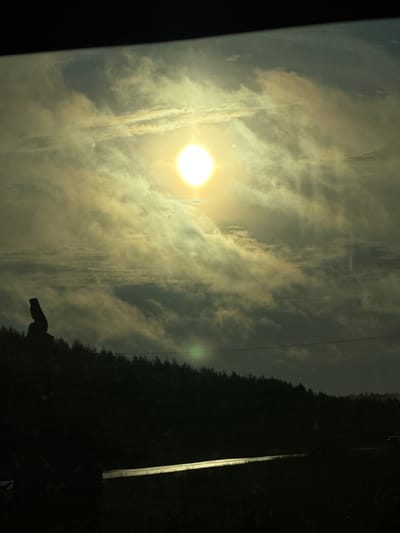 The image depicts a silhouetted figure standing on a dark, tree-lined hill against a dramatic, cloudy sky illuminated by a bright sun. The figure appears to be a statue or monument, with a distinctive angular shape. Below the hill, a dark road or surface reflects the light, suggesting it might be wet or metallic. The overall scene is dark and atmospheric, with the sun's glare dominating the upper portion of the frame. The specific location in the city of Яган-Докья, Russia, is not discernible from the image itself.