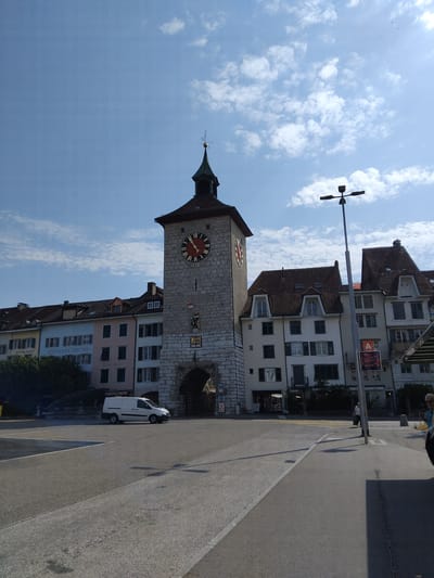 The image captures a scene in Solothurn, Switzerland, featuring the historic Baseltor (Basel Gate) tower prominently. The tower, constructed of stone, has a clock face with Roman numerals and red hands. Above the clock, a green-roofed spire is topped with a weather vane. To the left of the tower, a row of colorful buildings line the street, displaying architectural styles typical of the region. Some of these buildings appear to be shops or restaurants, with signs visible, such as "RESTAURANT" and "RESTAURANT BIELTOR". A white van is parked in front of one of the buildings.
To the right of the Baseltor, more buildings with dark tiled roofs and light-colored facades are visible. One building features a prominent orange sign with the letter "A". A modern street lamp with multiple light fixtures stands on the sidewalk. A woman in a light-colored top and dark skirt is walking on the sidewalk, carrying a bag. Further to the right, a glimpse of a person in a blue shirt is visible. The foreground consists of a paved road and sidewalk, with shadows cast by the buildings and the street lamp. The sky is a clear blue with scattered white clouds. The overall scene depicts a sunny day in a European city, with a blend of historic architecture and modern elements.