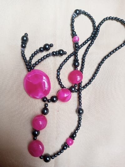 The image displays a single necklace arranged on a light tan fabric surface in Qarabucaq qəsəbə, Azerbaijan. No individuals are present. The necklace features a prominent color scheme of dark gray/black and hot pink. It is constructed from numerous small, spherical dark gray/black beads forming the primary strands, interconnected with larger, smooth, dark gray/black spherical beads and several hot pink beads. The hot pink beads vary in size and shape, including one large, irregular flat disc-like bead with white swirling patterns, positioned on the left. Other hot pink beads are spherical or disc-shaped, also exhibiting a marbled appearance. Small, textured, possibly metallic, spacers or caps are intermittently visible, particularly near larger dark beads and at the ends of some bead segments. The necklace is laid out in a curvilinear, abstract composition across the entire frame.