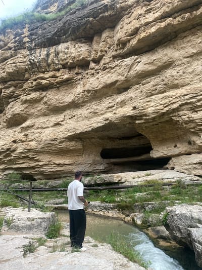 The image shows a man standing on a rocky bank of a fast-flowing river. He is wearing a white t-shirt and black pants. Behind him, a sheer cliff face rises, with a large cave opening visible in the rock. The environment is natural and rugged, with sparse vegetation growing on the rocks and cliff. The location is identified as Saltinsky Waterfall in the village of Saltá, Russia. The waterfall itself is not fully visible, but the powerful flow of water suggests a significant cascade.