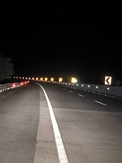An eye-level, full shot captures a highway at night with a white line dividing two lanes of asphalt. The lane on the left is bordered by a metal guardrail with several red reflectors. Along the right side of the highway, a series of elevated, yellow rectangular signs with black arrows indicate a curve to the left, interspersed with small, white reflective markers. The sky above is completely black. The scene appears to be located on a highway in the vicinity of Keureuweungkrueng, Indonesia.