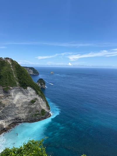 The image shows an ocean view from the cliffside of Nusa Penida, an island southeast of Bali, Indonesia, in the village of Bunga Mekar. The sky is blue with streaks of cirrus clouds. The ocean is a gradient of dark blue in the distance to turquoise near the shore. Several small, rocky islands rise from the sea. A small boat is visible on the water. A high cliff face covered with green vegetation occupies the foreground. A narrow trail is visible on the cliffside. The bottom right corner of the frame contains some green leaves.