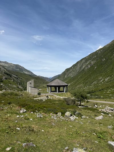 The image depicts a scenic mountain landscape in Hospental, Switzerland. In the foreground, a grassy slope is dotted with large rocks and sparse vegetation. A paved road runs through the middle ground, bordered by more rocks and low-lying green plants. Two prominent structures are visible: on the left, a modern, angular concrete building with a prominent vertical antenna, and on the right, a circular, open-sided pavilion with a dark, conical roof supported by pillars. Both structures are situated in a valley surrounded by steep, verdant mountains. The sky above is a bright blue, with scattered wispy clouds. The overall scene is one of natural beauty with architectural elements integrated into the environment.
