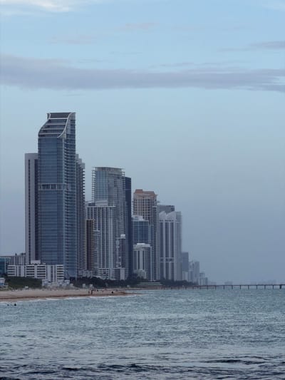 This is a vertical, eye-level, medium-range photograph of a beachfront skyline in Bal Harbour Village, United States of America, on an overcast day. The ocean occupies the foreground and lower portion of the image, with gentle waves lapping at a sandy beach. The beach is lined with a row of modern, high-rise buildings of varying heights and architectural styles. The buildings are predominantly made of glass and concrete, with some featuring balconies and patterned facades. A long pier extends from the right side of the frame out into the ocean, with silhouetted figures visible on it. A few scattered individuals are visible on the beach, and one person is swimming in the ocean. The sky above is a pale blue, with soft, elongated clouds. The overall mood is tranquil and slightly muted due to the overcast conditions.