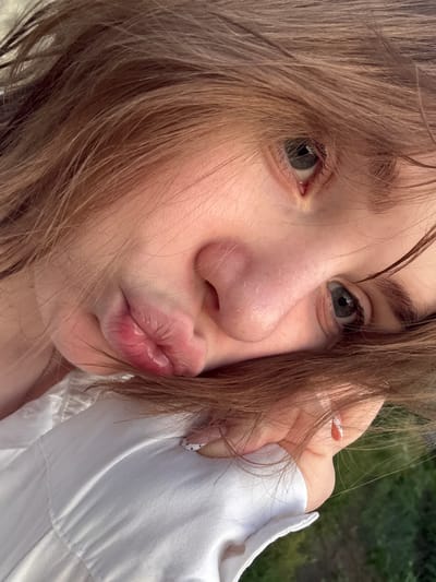 The image shows a close-up, angled view of a young person's face. Their brown hair is loose and frames their face, with strands falling over their eyes and cheek. The person's eyes are open and looking towards the camera, with dark lashes and brows. Their lips are slightly parted and appear plump, with a reddish hue. They are wearing a white garment with visible stitching and a small ring on their finger, which has a minimalist white and black polka dot design on one nail. The background is out of focus, showing hints of green foliage, suggesting an outdoor setting, possibly a park or garden. The lighting appears to be natural, with soft shadows. There is no information in the image to determine the location within the city of Ivatsevichy, Belarus.