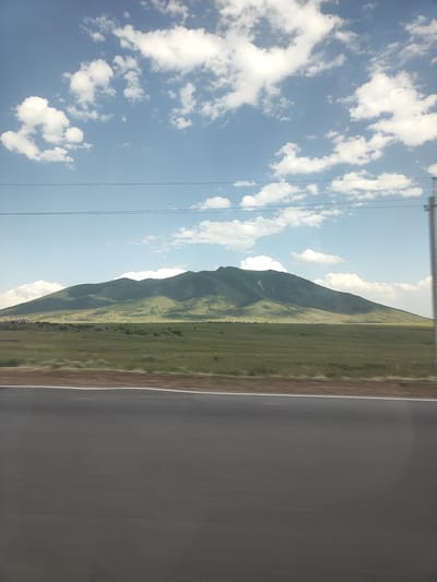 A wide, asphalt road with a white line marking runs horizontally across the lower third of the image. Beyond the road is a flat, grassy field with scattered rocks and vegetation. In the middle ground, a large, verdant mountain range dominates the landscape, with rolling hills and gentle slopes. The mountain peaks are covered in green grass, with some patches of snow or lighter colored soil visible near the summit. The sky above is a clear blue, dotted with fluffy white clouds. Overhead, two thin, dark power lines stretch horizontally across the scene. The image appears to have been taken from a moving vehicle, as evidenced by the slight blur of the road. The scene depicts a natural landscape with signs of human infrastructure. The location is specified as Apnagyugh, Armenia.