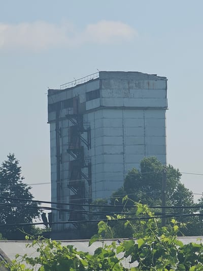 An aging, multi-story concrete structure with an exterior metal staircase stands against a hazy blue sky. The building appears weathered, with visible seams between the concrete panels and some signs of rust on the staircase. Trees and power lines are in the foreground and surrounding the base of the building, obscuring the ground level. The top of the building has a partially collapsed or damaged roofline. The overall scene suggests an industrial or abandoned site in Georgievka, Russia.