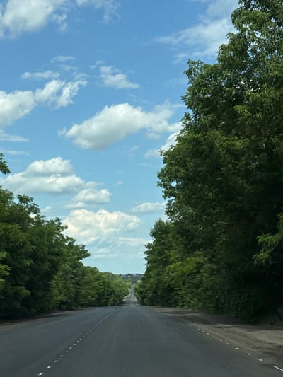 This media file depicts a road lined with dense green trees on either side, under a bright blue sky with scattered white clouds. The perspective is from a vehicle traveling down the road, suggesting movement. The road itself is asphalt and has a white dashed line marking its edge. In the distance, there appear to be a few small vehicles on the road, and a hint of buildings can be seen through the trees on the left side. The overall impression is that of a scenic drive through a rural or semi-rural area on a clear day. The time of day appears to be daytime, likely late morning or afternoon, given the bright sunlight. The weather is fair and pleasant. While specific emotions are not evident, the scene evokes a sense of peaceful travel or a scenic journey. No visible text is present in the image. The provided location context is Fursy, Ukraine, which aligns with the appearance of a country road.