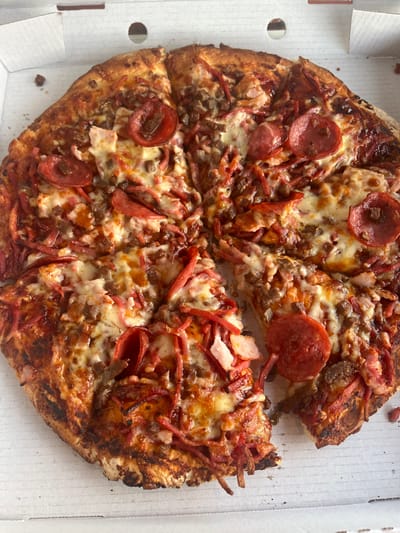 A round pizza, approximately 12-14 inches in diameter, is presented in an open, white corrugated cardboard pizza box. The pizza is sliced into multiple pieces, with one slice partially removed or missing from the bottom right quadrant. The crust exhibits a golden-brown coloration with localized areas of charring. Toppings consist of melted white cheese, numerous circular slices of pepperoni, crumbled brown ground meat, and reddish-pink shredded meat strips. A small, dark crumb is visible in the upper left corner of the pizza box. The box features several small, circular ventilation holes along its upper perimeter. The scene is situated in the city of Applecross, Australia. No individuals, actions, or direct interactions are visible in the image.