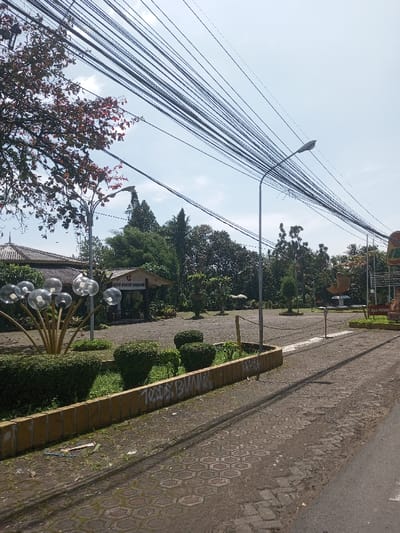 This image depicts an outdoor public area, possibly a park entrance or plaza, in Beteng_Pangkhar_Somthrang, Bhutan, under a clear, bright sky with scattered clouds. Numerous black utility wires are strung horizontally across the upper portion of the frame.
The foreground features a dark grey paved road or curb transitioning to a hexagonal-patterned grey paver walkway. Along the walkway, low garden beds are defined by a yellow-bricked retaining wall, which has light-colored graffiti including "TODS BUNT" visible. These beds contain manicured green bushes and grass. A light-colored sandal and other debris are visible on the ground near the wall.
In the mid-ground, a decorative sculpture resembling clear spherical bubbles on golden-colored stems stands within a garden bed to the left. Behind it, a building with a dark, multi-tiered roof is partially visible. Further into the scene, an open, grey paved area, likely a parking lot or plaza, stretches towards the background. A small, open-sided wooden pavilion with a light-colored roof and a sign bearing the text "DAN PAHAR SEMAR" is centrally located within this paved expanse.
Two grey metallic street light poles are visible; one on the far left, partially obscured by a tree with reddish-brown leaves, and another prominent pole extending upwards from the paved area to the right of center, intersecting the overhead wires. To the right of this central light pole, a short wooden post with a chain attached defines a boundary. An abstract, light-colored sculpture with curved forms is situated in the paved area to the mid-right. A large, colorful sign with text including "KHA PREMAYA" and "BUK" is partially visible on the far right. The background consists of dense green foliage from various trees and shrubs, creating a park-like environment. No individuals are present in the scene, and no discernible actions are occurring.