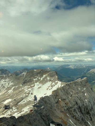 The image depicts a panoramic view of a mountainous region, likely taken from a high vantage point. Jagged, rocky peaks dominate the foreground and midground, with patches of snow scattered across their slopes. In the distance, a vast expanse of layered mountains fades into a hazy blue horizon. The sky is filled with a dramatic arrangement of clouds, ranging from dense, grey masses to thinner, wispy formations that allow glimpses of blue sky.

Specific structures are visible on the mountainside. To the left, a complex of buildings, possibly a resort or research facility, is nestled into the terrain. A prominent metal tower with a dish antenna stands on a rocky outcrop in the mid-right of the frame, suggesting a communication or observation post. The ruggedness of the mountains suggests a high-altitude, alpine environment. While the image is attributed to Ehrwald, Austria, specific landmarks within Ehrwald itself are not definitively identifiable from this perspective. The scene is devoid of any visible human figures or active events.