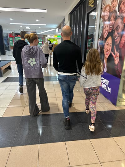 A group of four people are walking through a shopping center. The group consists of two adults and two children. The two adults appear to be a man and a woman, and the two children are also a boy and a girl. They are all walking away from the camera, and their faces are not visible. The shopping center appears to be indoors, with a tiled floor and fluorescent lighting. There are shops visible in the background, including a Subway and a Robin Hood. A large poster of smiling faces is also visible on the right side of the image. The people are dressed in casual clothing. The man in the foreground is wearing a black t-shirt and blue jeans. The woman is wearing a purple hoodie and grey pants. The boy is wearing a black hoodie and blue jeans. The girl is wearing a white and black patterned sweater and floral leggings. They are walking on a section of the floor that is tiled with a darker shade of tile, bordered by a lighter shade of tile. The context provided by the poster and the store names suggests this location is likely toormina, Australia.