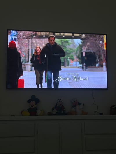 This vertically oriented image captures a flat-screen television displaying what appears to be the end credits of a film or television show. The TV screen itself is rotated 90 degrees counter-clockwise relative to the frame.

On the television screen, the prominent text reads "music by Justin Melland" in an elegant, script-like font. Below and to the left of the text, an outdoor scene is depicted. Three individuals are visible on what appears to be a city street or sidewalk. A man in a dark jacket and blue jeans stands facing towards the left side of the screen. Behind him, a woman with long, reddish-brown hair, wearing a dark top and lighter trousers, is smiling. In the foreground, partially obscured, is a person seen from the back, wearing a dark jacket and a red hat or hood, carrying a red bag. The background shows bare trees, suggesting a cool season like autumn or winter, along with hints of urban structures.

To the right of the television, several plush toys are arranged on a surface, likely a wall shelf or the top of a piece of furniture. These include a Mickey Mouse character plush, a Santa Claus-themed doll, and other lighter-colored stuffed animals. Further to the right, a white, two-paneled cabinet door is partially visible. The room where the photograph was taken appears dimly lit, which accentuates the brightness of the television screen.