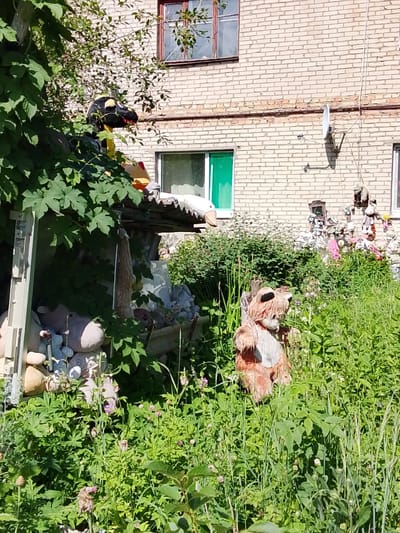 In a sunlit outdoor area adjacent to a pale brick building in Снопок Новый, Russia, a collection of stuffed toys and garden decorations are partially obscured by lush green foliage. A black and yellow dragon head toy is visible on the left, peeking over a leafy overhang. To the right, a larger, light brown plush fox-like toy stands upright amidst tall grass and small pink flowers. The background features the brick building with a green-trimmed window and a satellite dish mounted on the wall. Various other stuffed animals and objects are scattered behind the fox, some hanging from the building or resting amongst the vegetation, creating a scene of an overgrown, decorated garden.