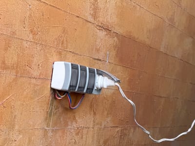 This is a close-up, slightly angled shot of a white electrical cord plugged into a gray and white junction box mounted on a textured, orange-brown wall. The wall appears to be made of horizontal planks, with visible seams and a rough, uneven surface that suggests it might be stucco or a similar material. The electrical cord is white and twisted, leading away from the junction box towards the top left of the frame. The junction box has several dark gray sections and a white end. Wires of red, blue, and possibly pink are visible protruding from the junction box. The lighting suggests an indoor setting, possibly with artificial light, as there are no strong shadows or indications of natural light sources. There is no visible text in the image. The scene appears to be a functional, practical setup, possibly for electrical connections, and does not convey any particular emotion or specific time of day beyond being indoors.